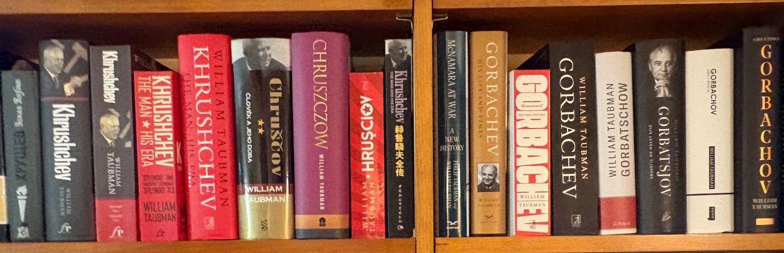 A shelf of William Taubman's books on Khrushchev and Gorbachev, hardback and paperback, including original English editions and translated editions in several languages. Predominant colors of the book covers are red and black, with some purple, gold, white, and tan. The books are framed by edges of light wooden shelves at top and bottom, and a wooden standard in the center.