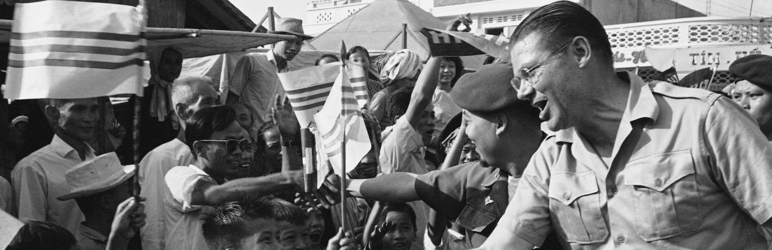 McNamara, in safari shirt, is at right side of photo, with his right hand reaching downward, where the tops of Vietnamese children's heads can be seen. One of the children holds a small flag displaying three horizontal stripes against a white background. Behind McNamara's right shoulder the head and extended right arm of a uniformed Vietnamese soldier can be seen, his head facing sidewards, towards a small crowd of Vietnamese people, some holding similar flags.