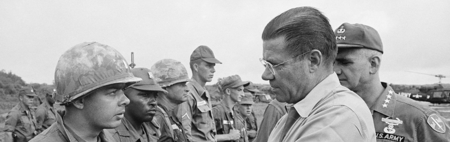 McNamara, head and shoulders in profile at right of photo, wearing dress shirt and tie, with an officer partially visible behind him in field uniform with "US Army" and four stars on his shirt. At the left and facing forward are several soldiers in fatigues, two in camouflage-covered helmets and the others in caps. The soldier in the foreground appears to be in conversation with McNamara. A helicopter is visible in the distant background.